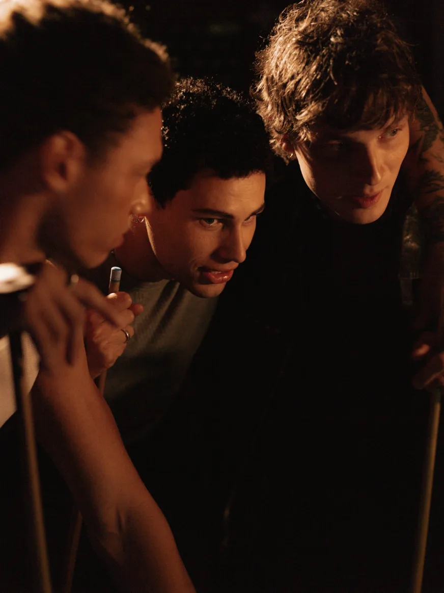 Three young men with full hair gathered together in a warm-lit room