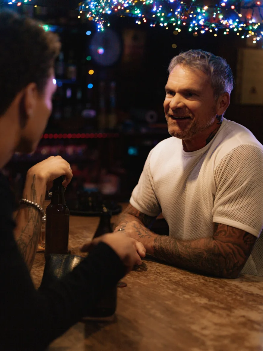 Older man with tattooed arms smiling at a friend across a bar counter under string lights