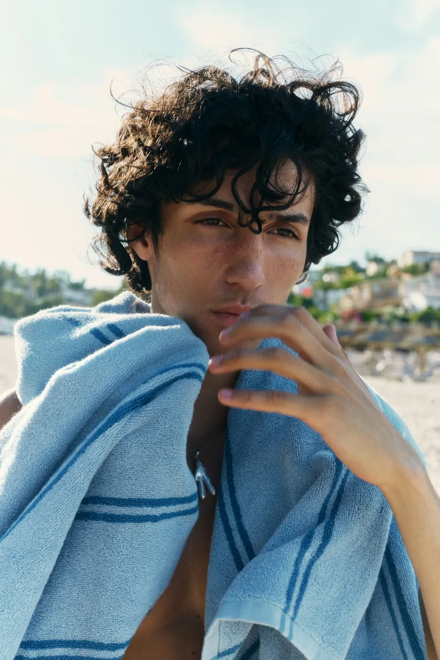 Young man with thick curly dark hair holding a striped towel at the beach